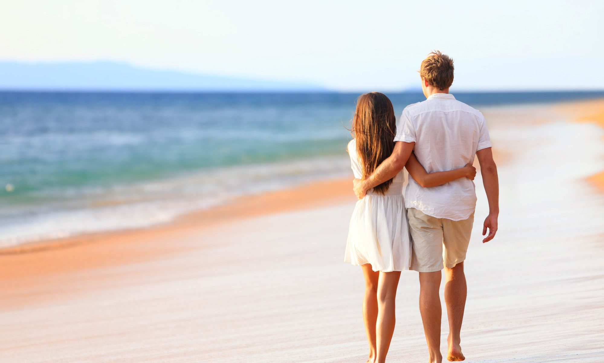 A couple sitting on a sandy beach at sunset, holding hands and smiling, with a beautiful colorful sky in the background.